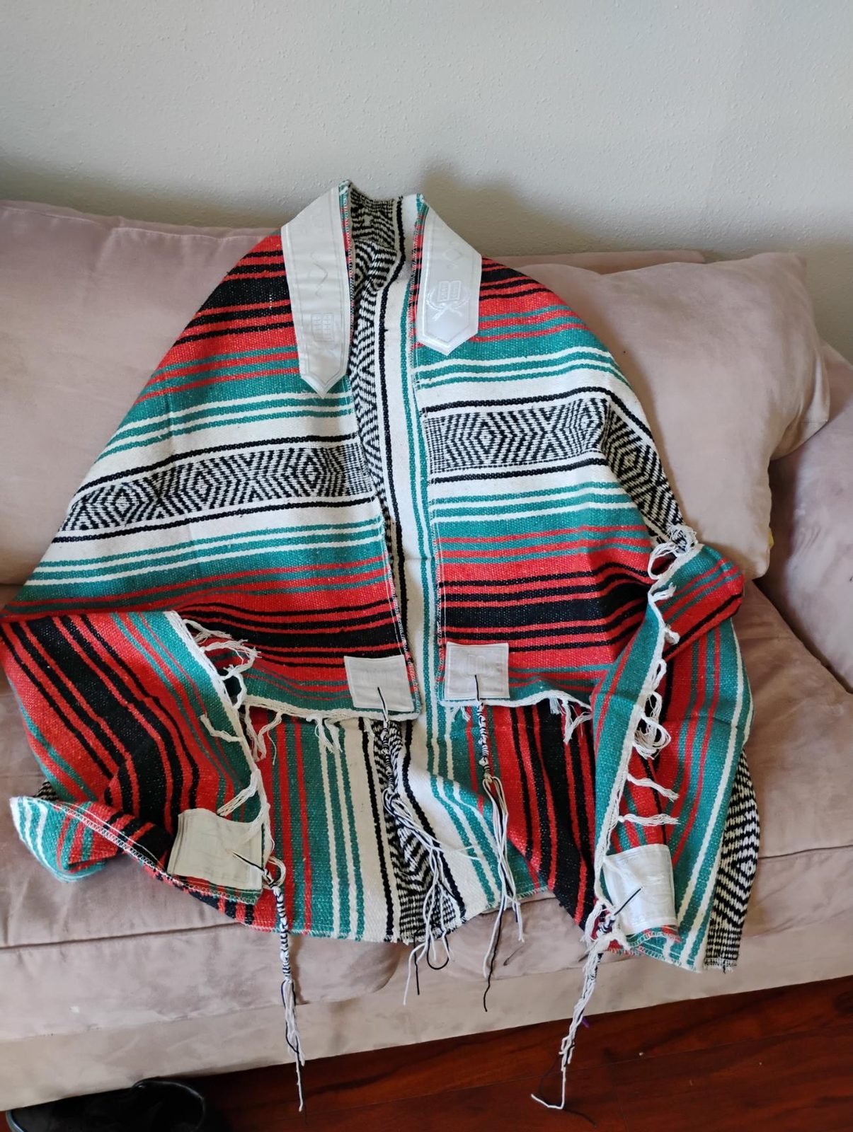 Turquesa sarape tallit spread on a sofa: teal, red, black, and white stripes, a white atarah at the neck, corner plates, and tzitzit.